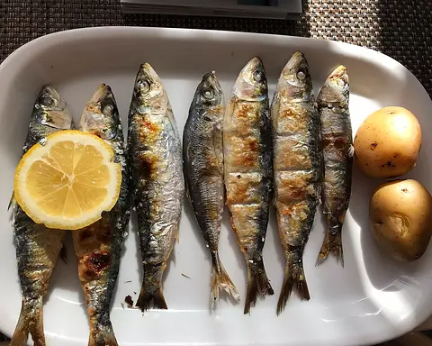 Seven grilled sardines on a white platter with a slice of lemon and two boiled potatoes